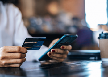 Close-up de uma mulher sentada em uma mesa de madeira, segurando um cartão de crédito na mão esquerda e um smartphone na direita, digitando os dados do cartão para realizar uma compra online. Ao fundo, um copo de café para viagem e um notebook fechado.