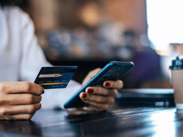 Close-up de uma mulher sentada em uma mesa de madeira, segurando um cartão de crédito na mão esquerda e um smartphone na direita, digitando os dados do cartão para realizar uma compra online. Ao fundo, um copo de café para viagem e um notebook fechado.