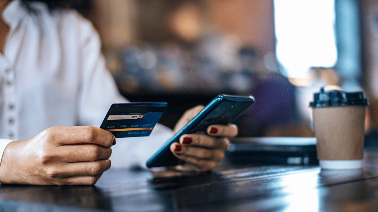 Close-up de uma mulher sentada em uma mesa de madeira, segurando um cartão de crédito na mão esquerda e um smartphone na direita, digitando os dados do cartão para realizar uma compra online. Ao fundo, um copo de café para viagem e um notebook fechado.